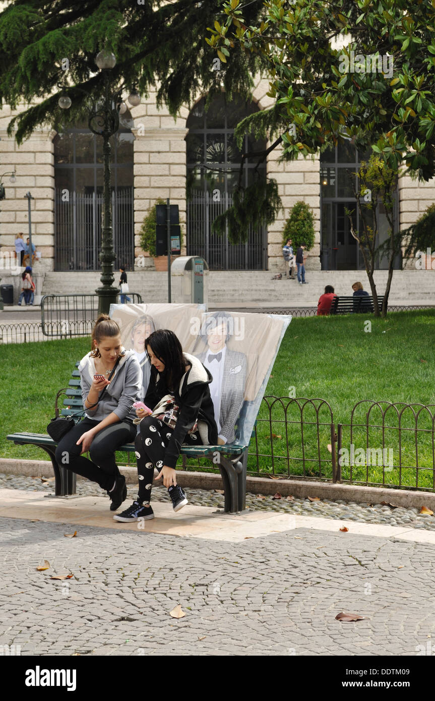 Zwei Mädchen im Teenageralter ruhen auf einer Parkbank in Piazza Bra, Verona, mit Postern von ihren Liebsten eine Richtung Bandmitglieder. Stockfoto