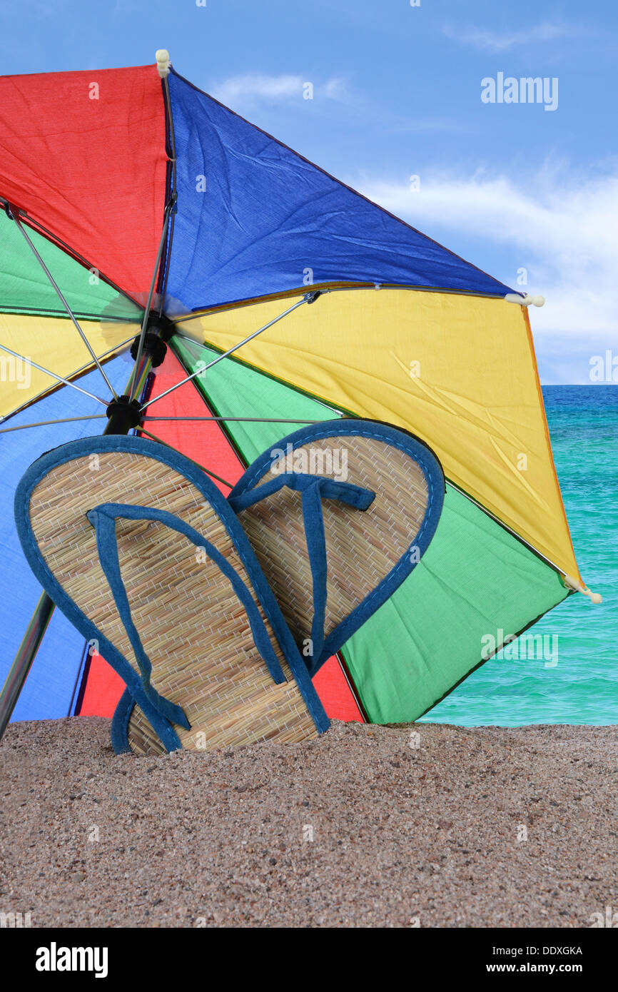 Nahaufnahme von ein paar Flip-flop-Sandalen sind mit einem bunten Sonnenschirm im Sand stecken. Stockfoto