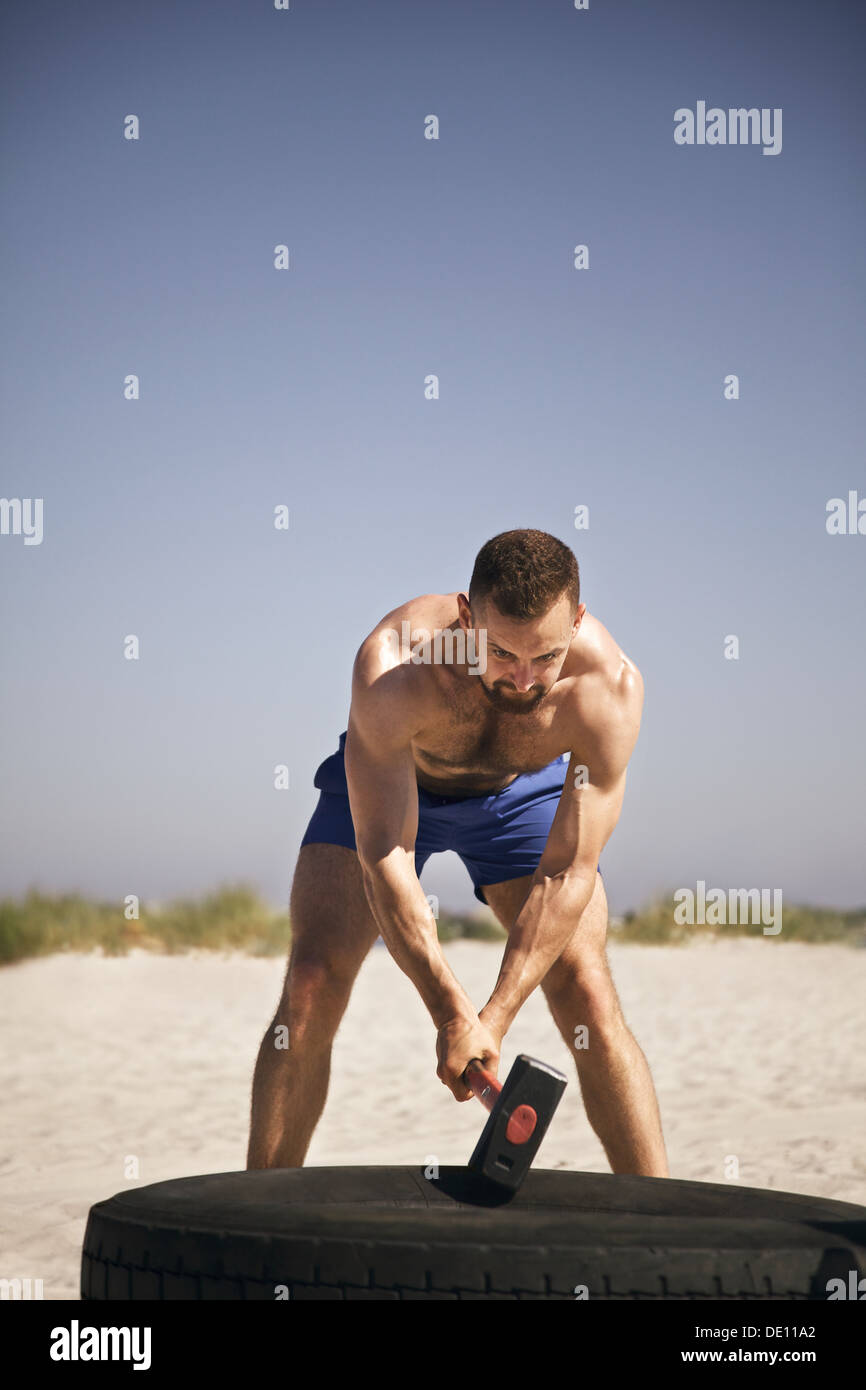 Männlicher Athlet Hämmern LKW-Reifen mit einem Vorschlaghammer beim Crossfit Training am Strand Stockfoto