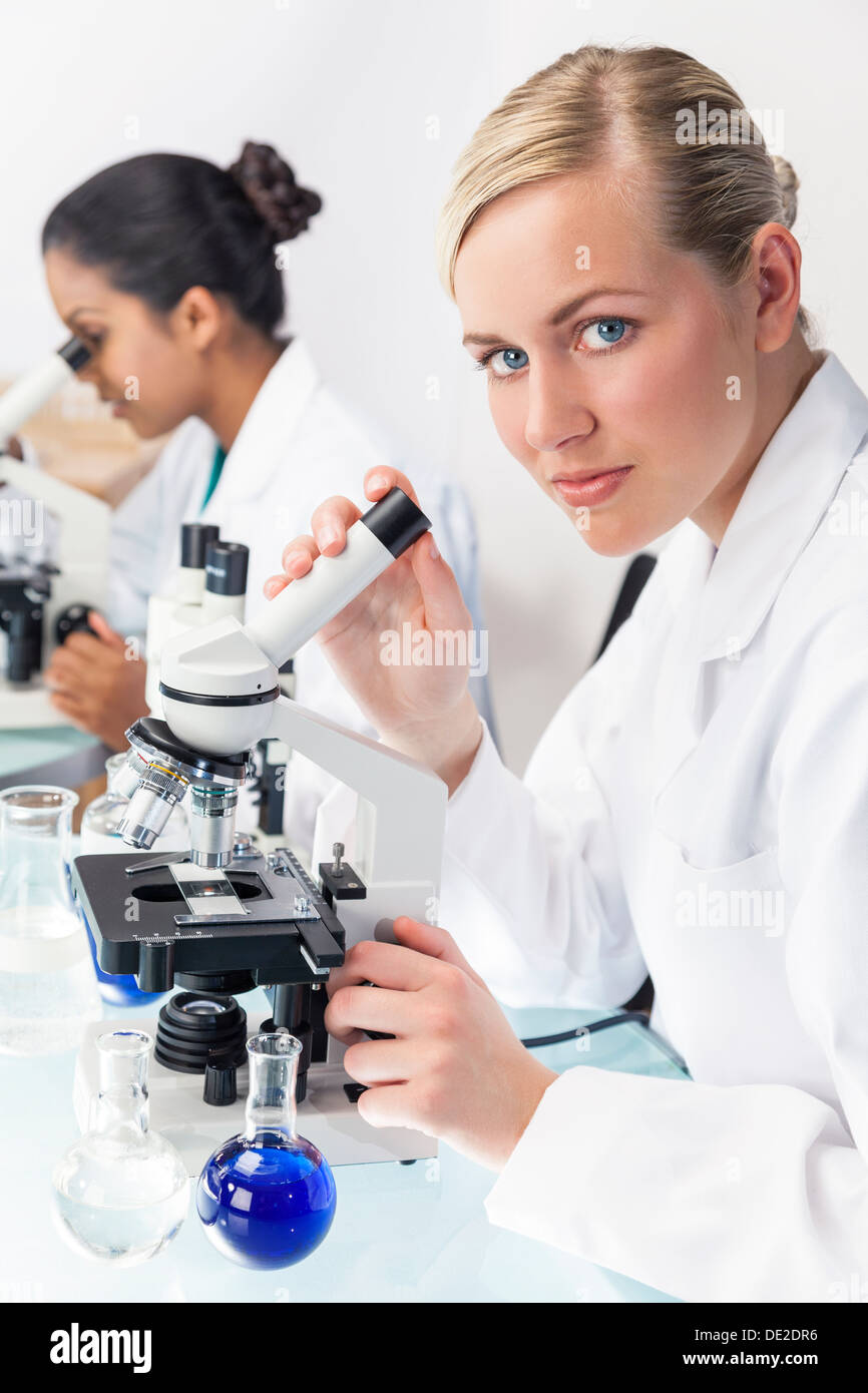 Ein blondes medizinische oder wissenschaftliche Forscherin oder Arzt mit ihrem Mikroskop im Labor mit ihrem asiatischen Kollegen. Stockfoto