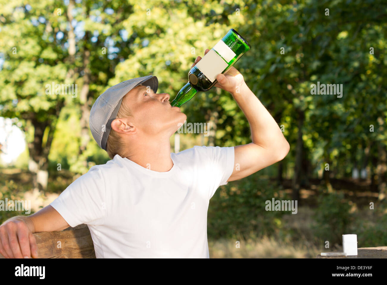 Männliche Täter Alkohol trinken aus einer Flasche Weißwein sitzen auf einer Bank im Park an einem warmen Sommertag Stockfoto