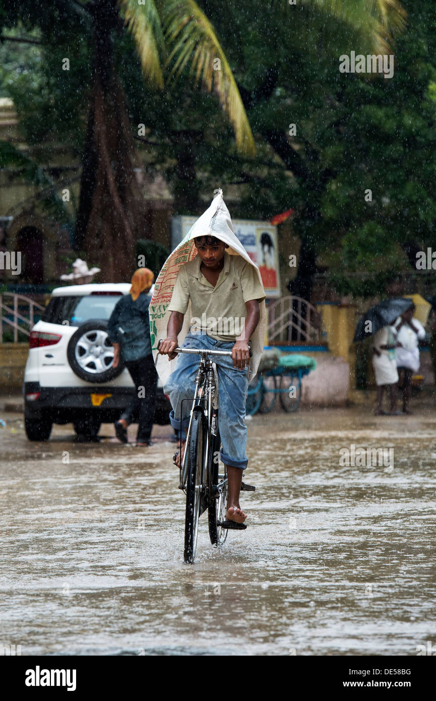 Indischer jugendlicher Radfahren durch eine überflutete Straße mit einer Tasche auf den Kopf. Puttaparthi, Andhra Pradesh, Indien Stockfoto
