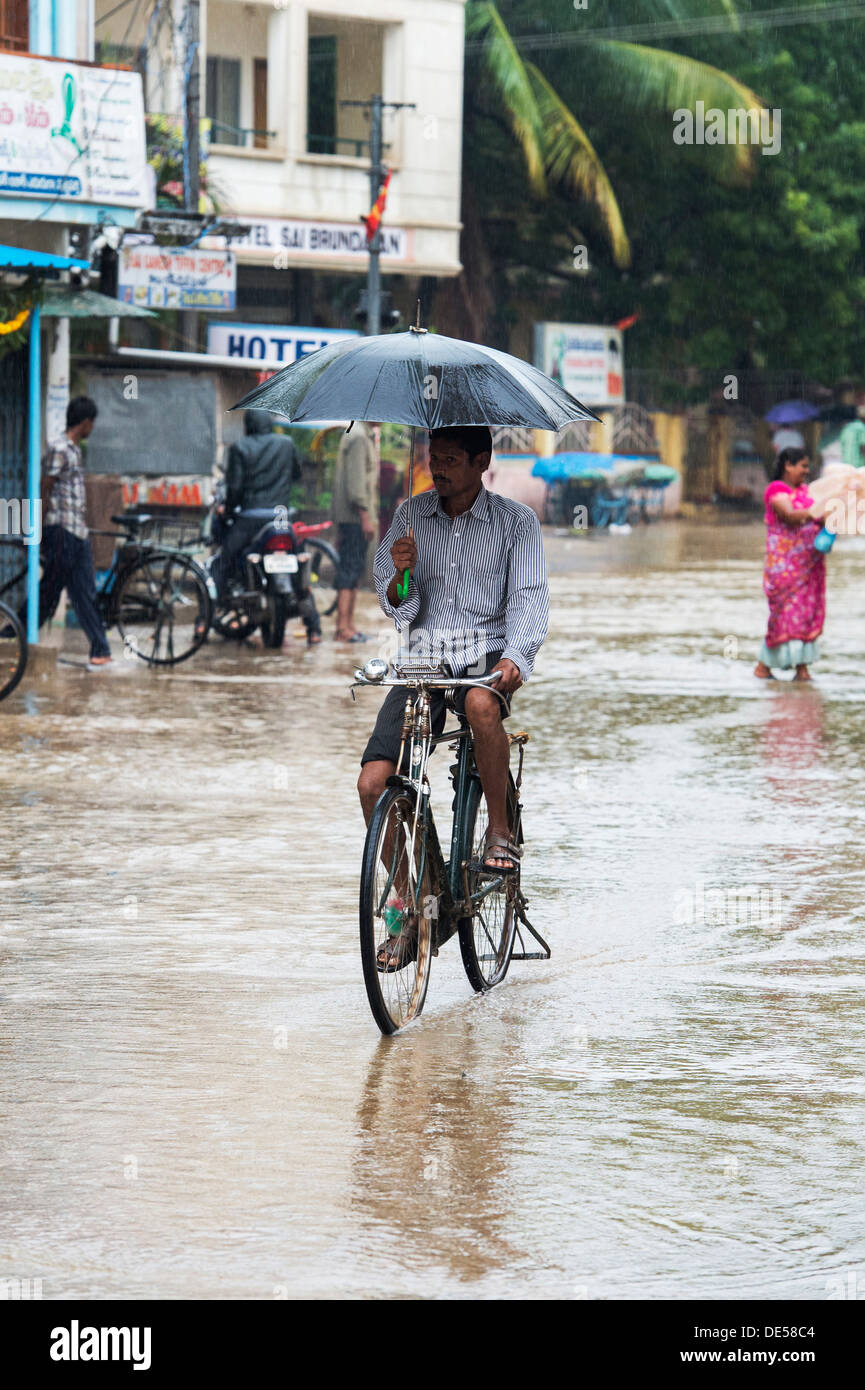 Indischer Mann Radfahren durch eine überschwemmte Straße tragen einen Regenschirm. Puttaparthi, Andhra Pradesh, Indien Stockfoto