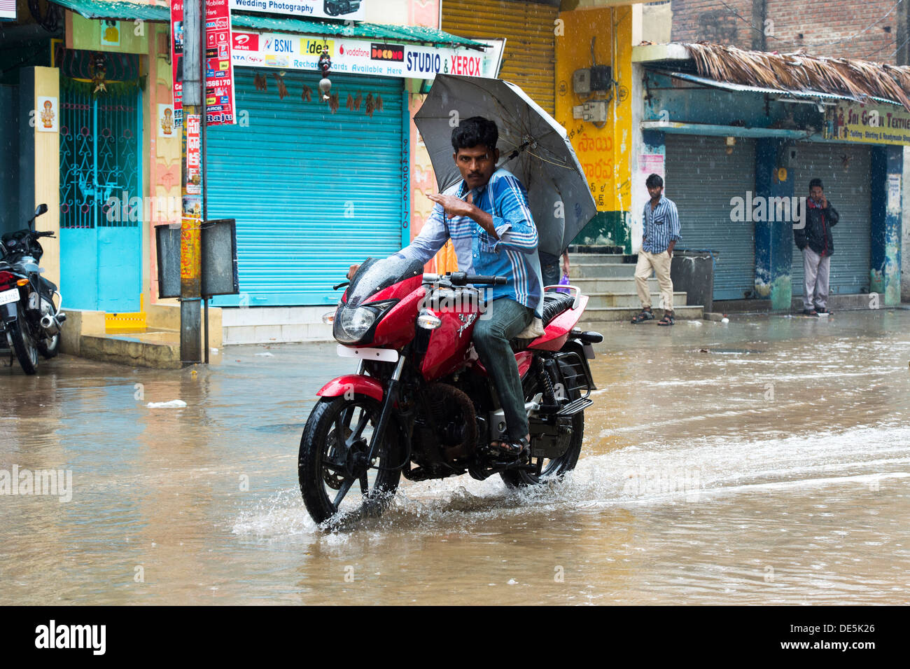 Indischer Mann mit einem Regenschirm auf seinem Motorrad reiten durch nasse Straßen in Puttaparthi, Andhra Pradesh, Indien Stockfoto