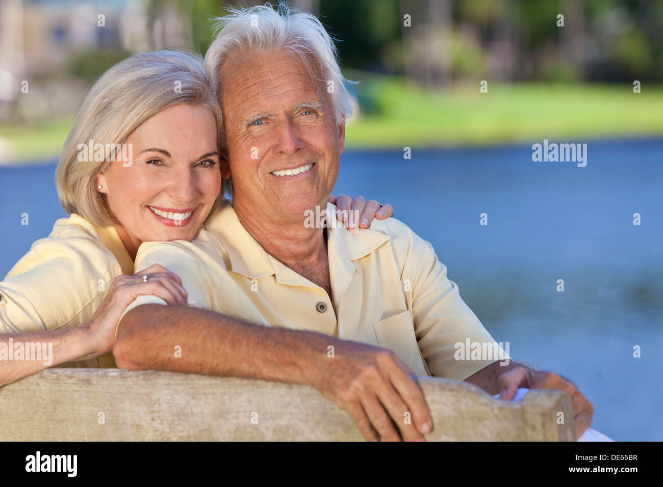 Glücklich lächelnd romantisch senior Mann und Frau zu zweit sitzen auf einer Parkbank, umarmt von einem blauen See Stockfoto