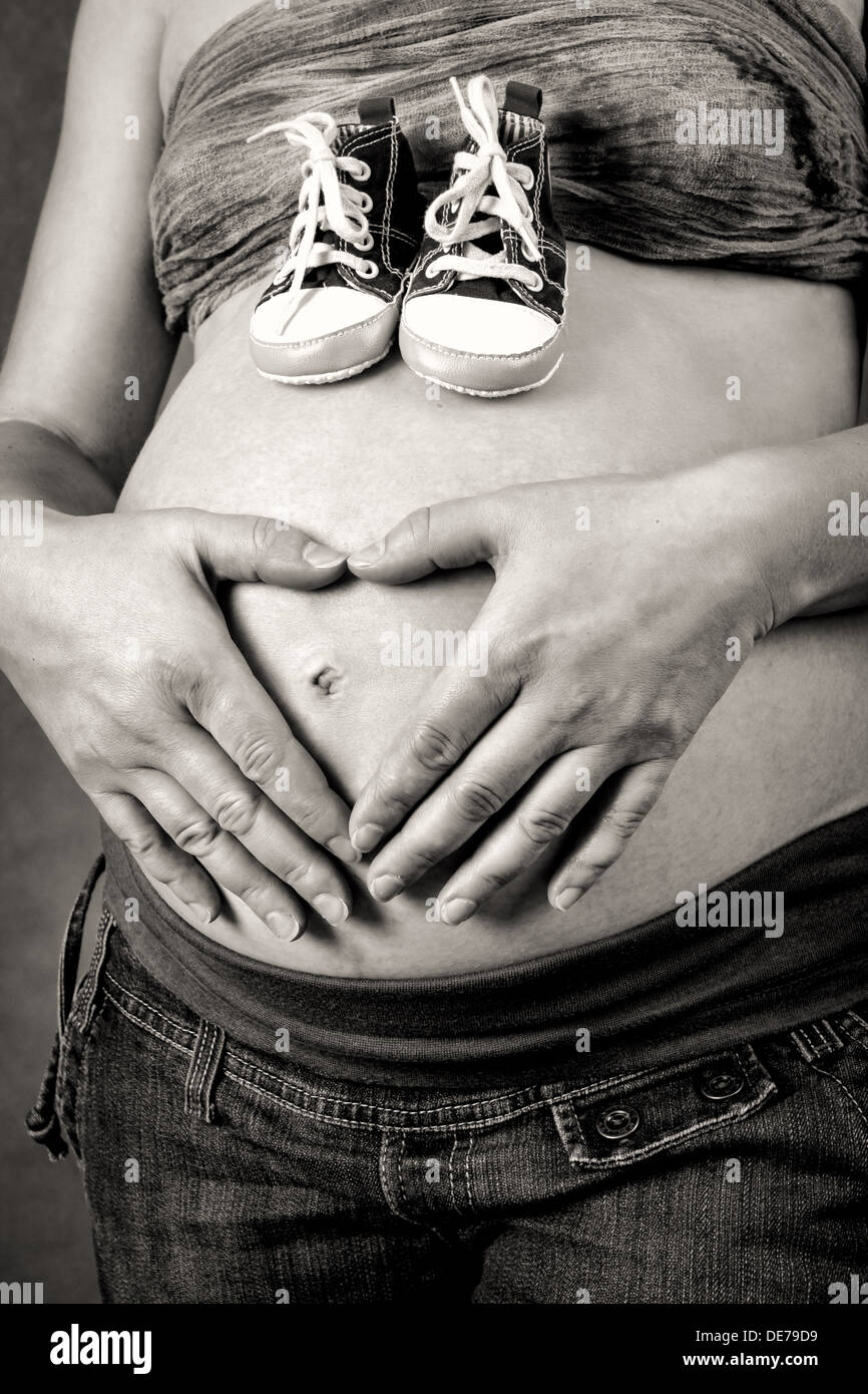 Schwangere Frau mit einem Kind Schuh zärtlich hält ihren Bauch mit der Hand als Herzform Stockfoto