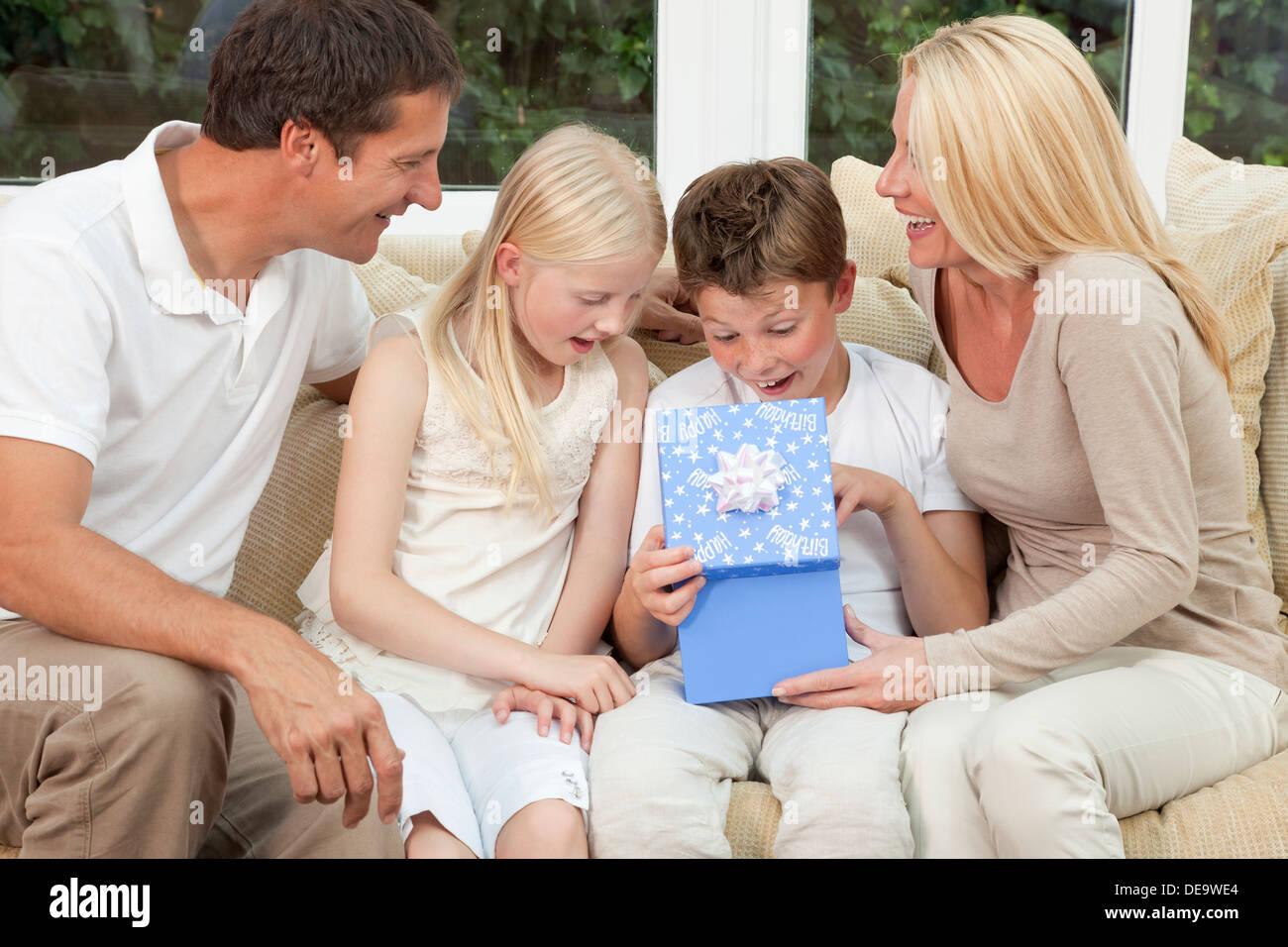 glückliche Familie Mutter Vater Sohn und Tochter sitzen auf einem Sofa zu Hause das junge Kind ist ein Geburtstagsgeschenk in einer blauen Schachtel öffnen Stockfoto