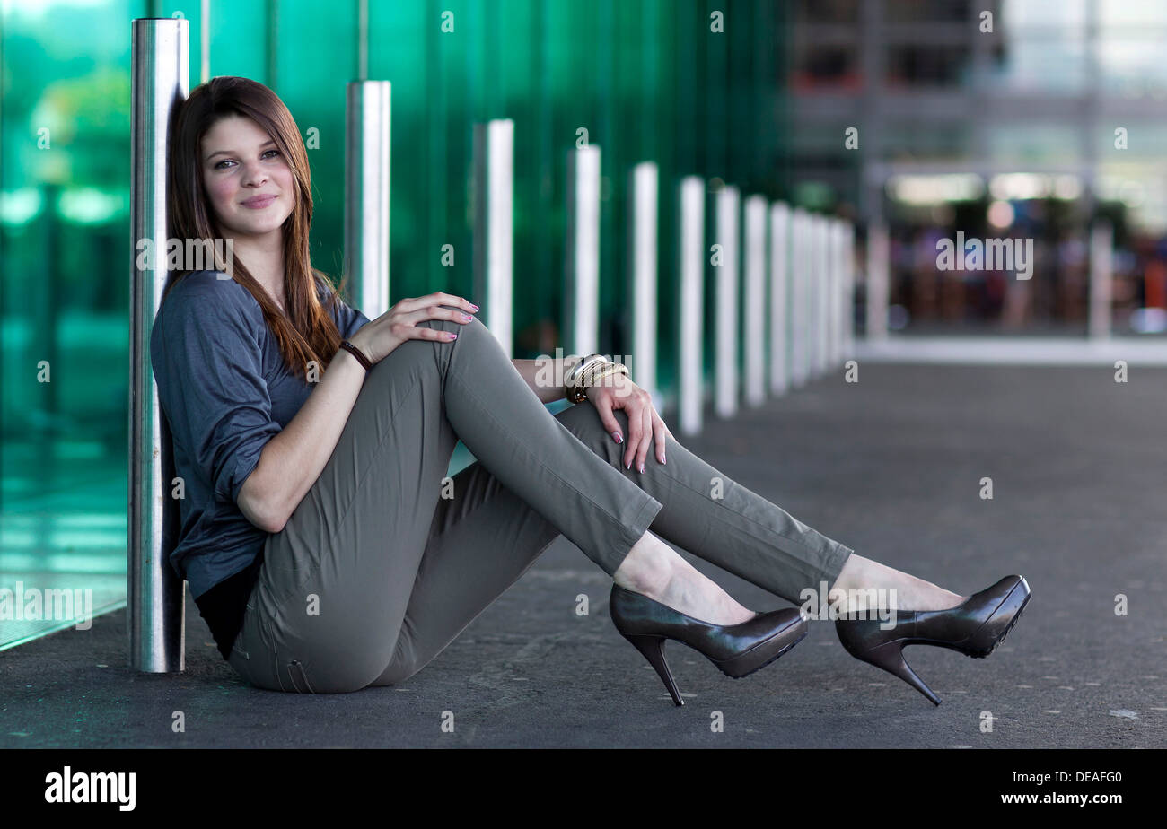 Lächelnde junge Frau mit high Heels sitzt und posiert vor einer grünen Glaswand Stockfoto