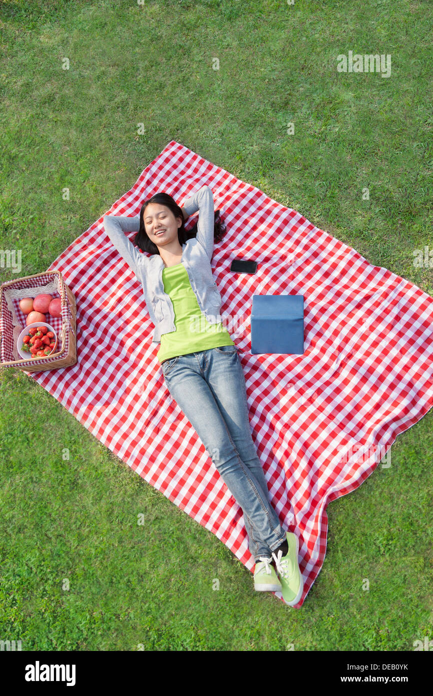 Lächelnde junge Frau mit Armen hinter ihrem Kopf auf einer Decke auf dem Rücken liegend und Relaxen im park Stockfoto