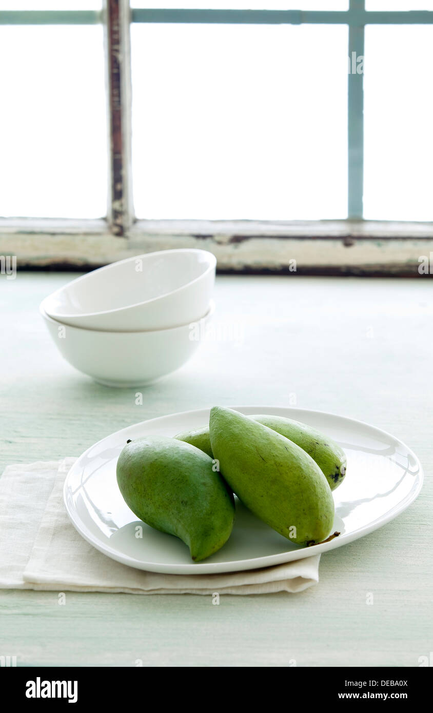 Ein Teller mit grünen Mangos neben weißen Schalen und einem sonnigen Fenster. Stockfoto