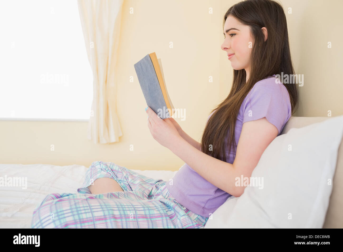 Lächelndes Mädchen sitzt auf einem Bett, ein Buch zu lesen Stockfoto
