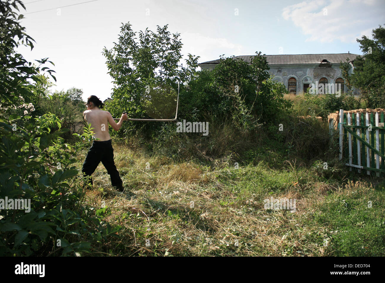 Mann, mähen den Rasen im Sommer. Plotinnoye Dorf, Krim, Ukraine. Hinter steht das verlassene Haus der Kultur. Stockfoto