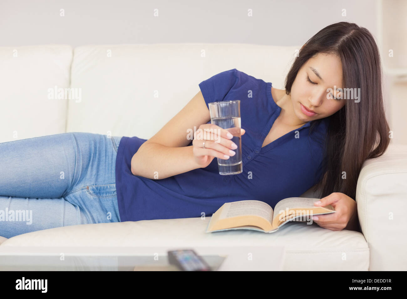Gerne Asiatin lag auf dem Sofa liest einen Roman und Trinkwasser Stockfoto