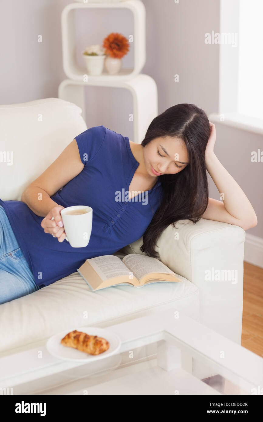 Hübsche junge Asiatin auf dem Sofa ein Buch hält ihren Kaffee Stockfoto