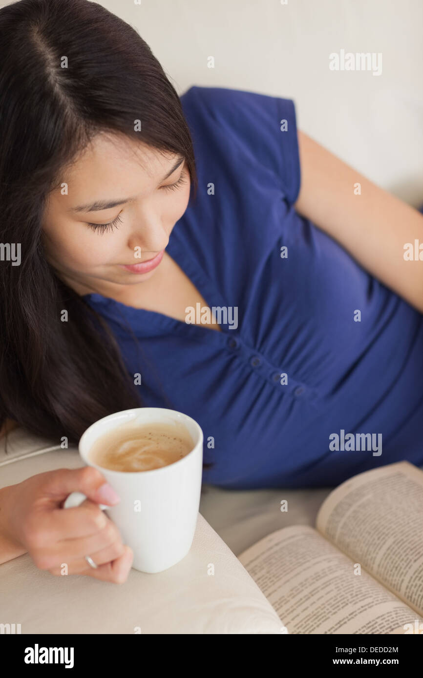 Junge asiatische Frau lag auf dem Sofa ein Buch hält ihren Kaffee Stockfoto