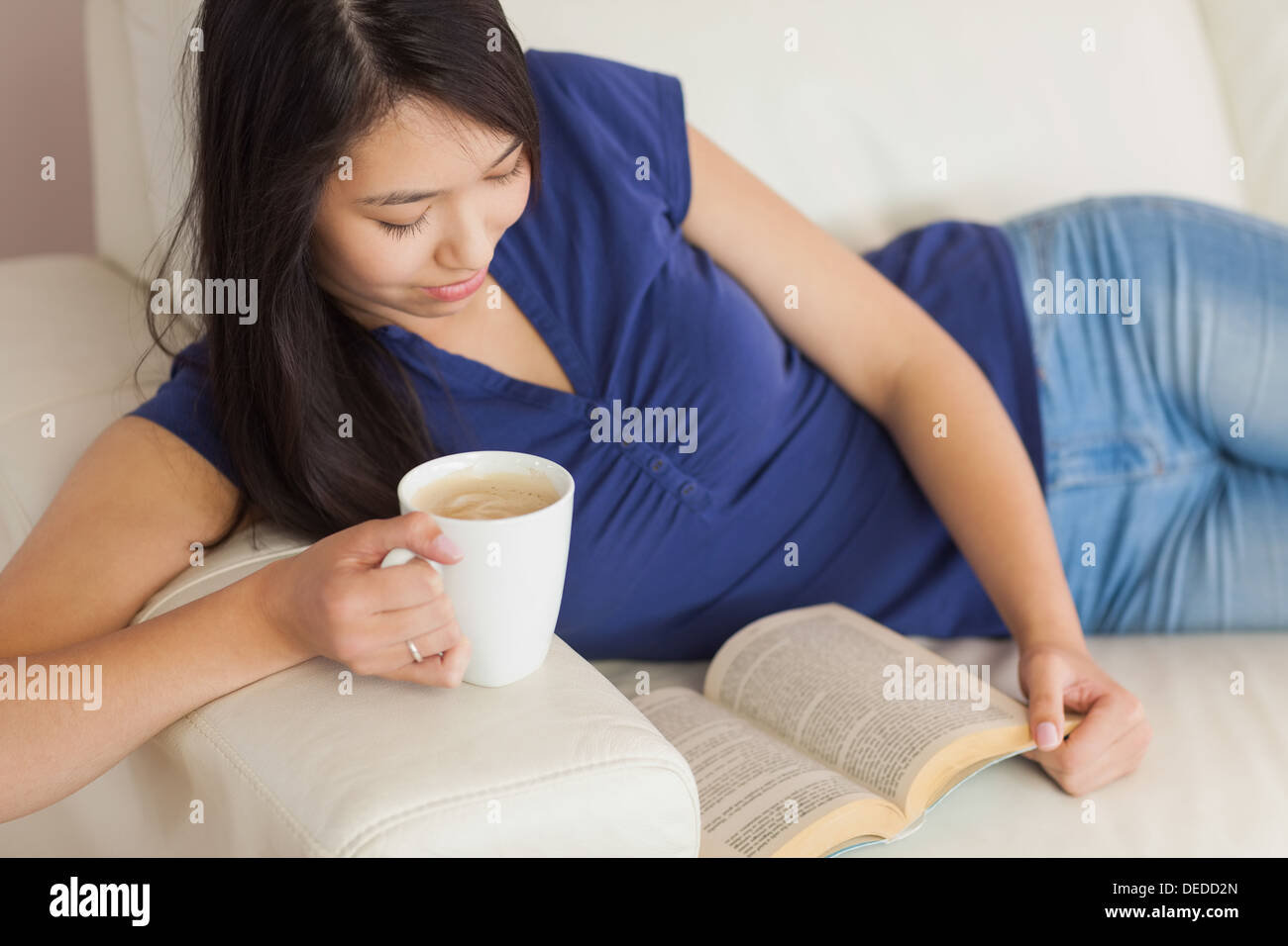 Glückliche junge asiatische Frau lag auf dem Sofa ein Buch hält ihren Kaffee Stockfoto