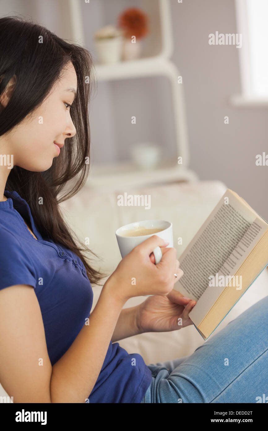 Junge asiatische Frau sitzt auf dem Sofa ein Buch hält ihren Kaffee Stockfoto