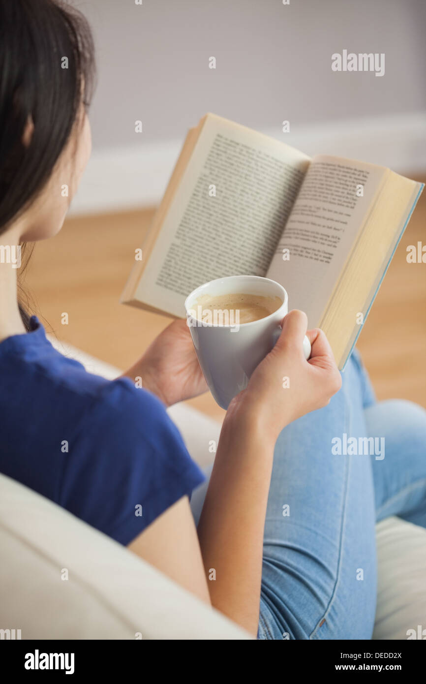 Junge Frau sitzt auf dem Sofa ein Buch hält ihren Kaffee Stockfoto