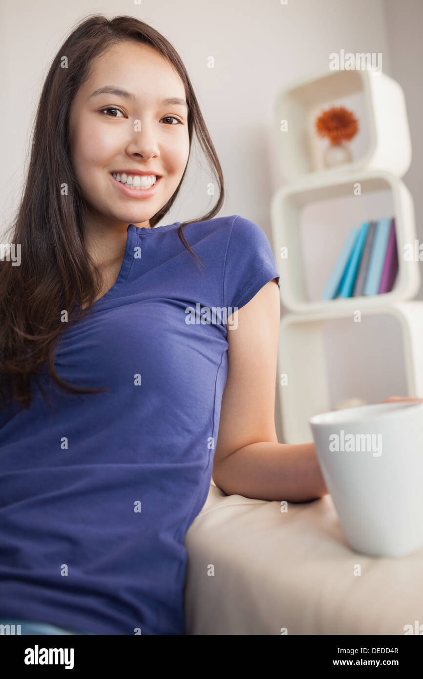 Glückliche junge asiatische Frau sitzen auf der Couch halten Becher Stockfoto