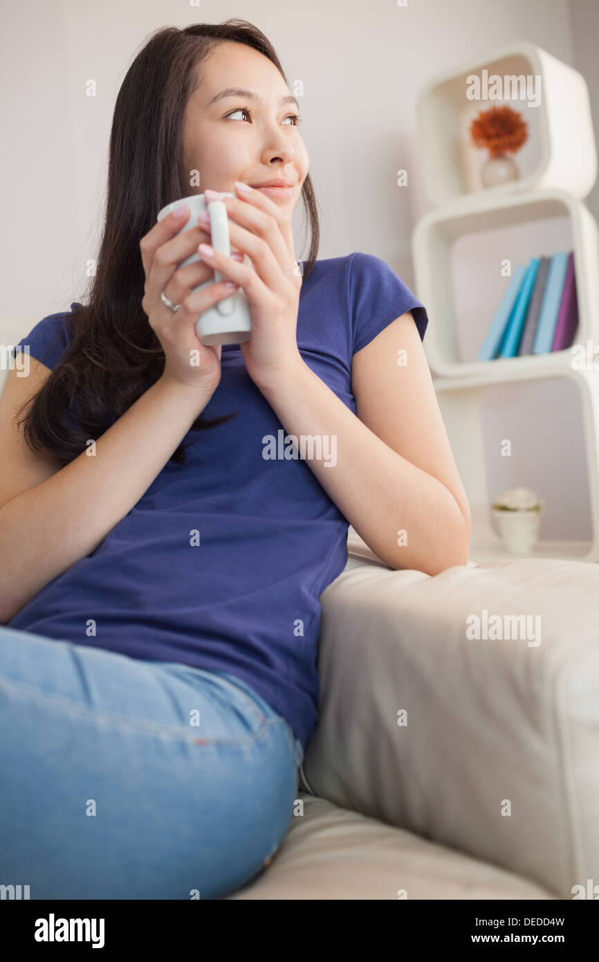 Nachdenkliche junge Asiatin auf der Couch halten Becher Stockfoto
