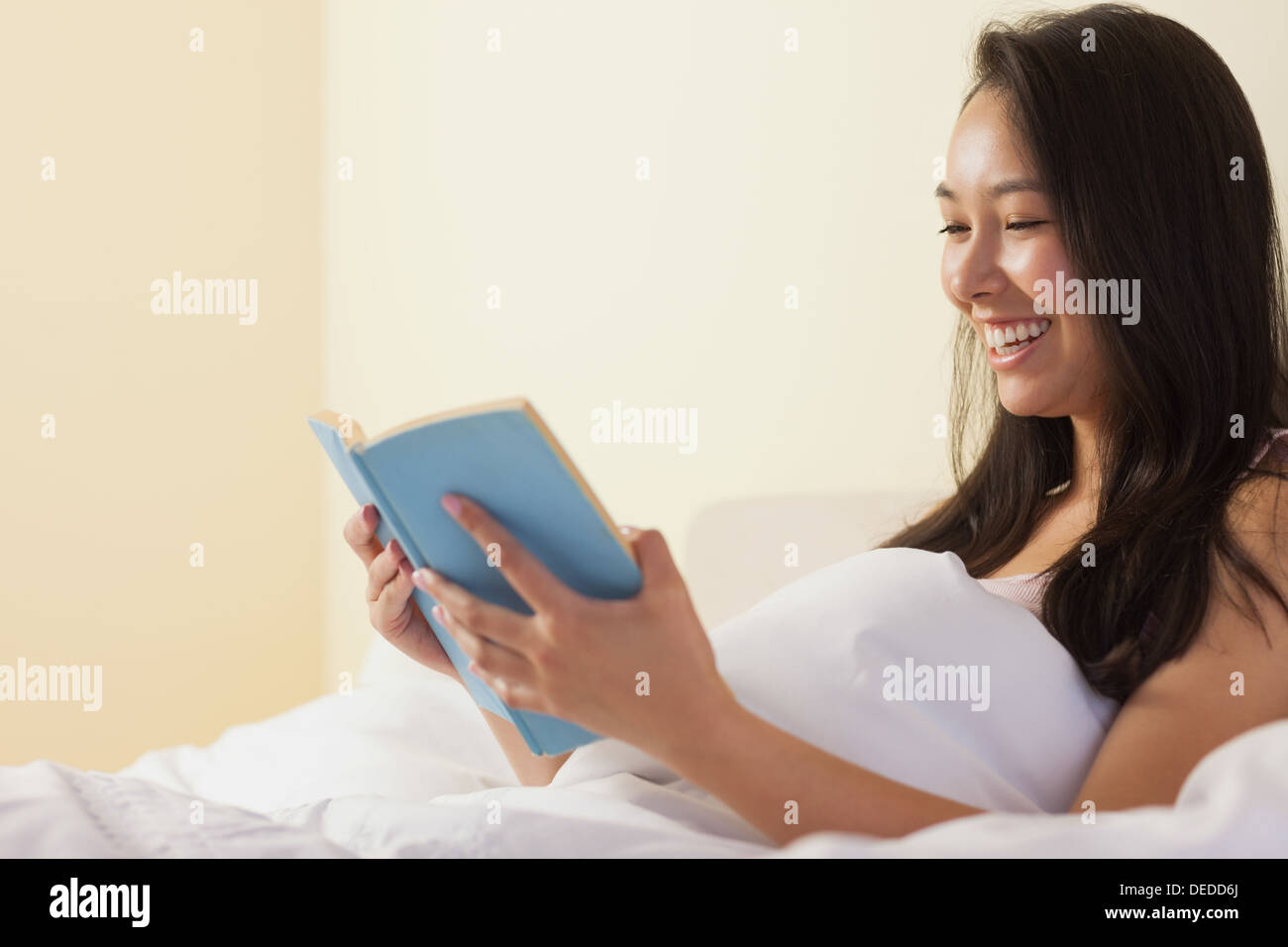 Lächelnde junge asiatische Frau sitzen im Bett und las ein Buch Stockfoto