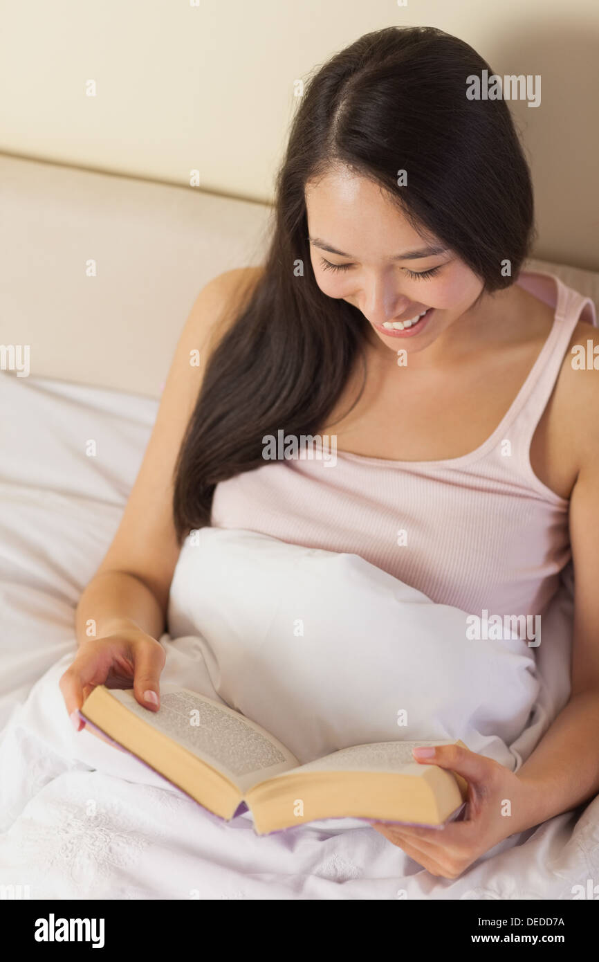 Fröhliche junge asiatische Frau sitzen im Bett und las ein Buch Stockfoto