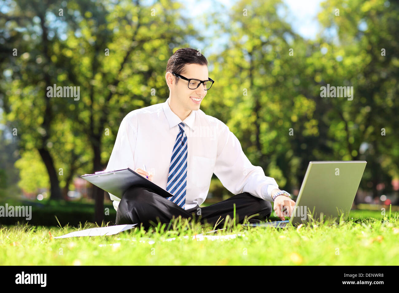 Junger Buchhalter sitzen auf einer Wiese auf einem Laptop arbeiten und aufschreiben von Notizen in einem park Stockfoto