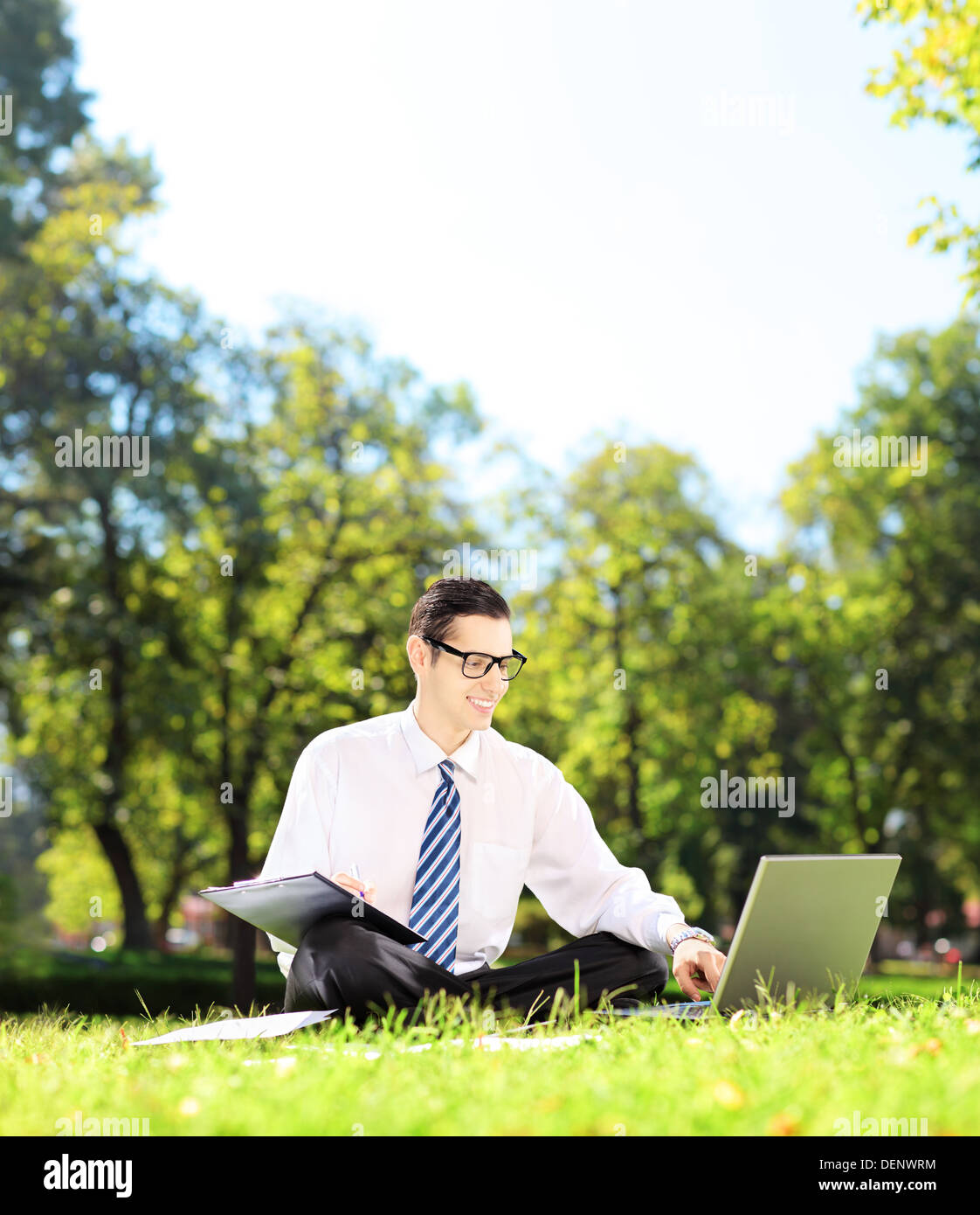 Junger Geschäftsmann sitzt auf einem grünen Rasen auf einem Laptop arbeiten und aufschreiben von Notizen in einem park Stockfoto