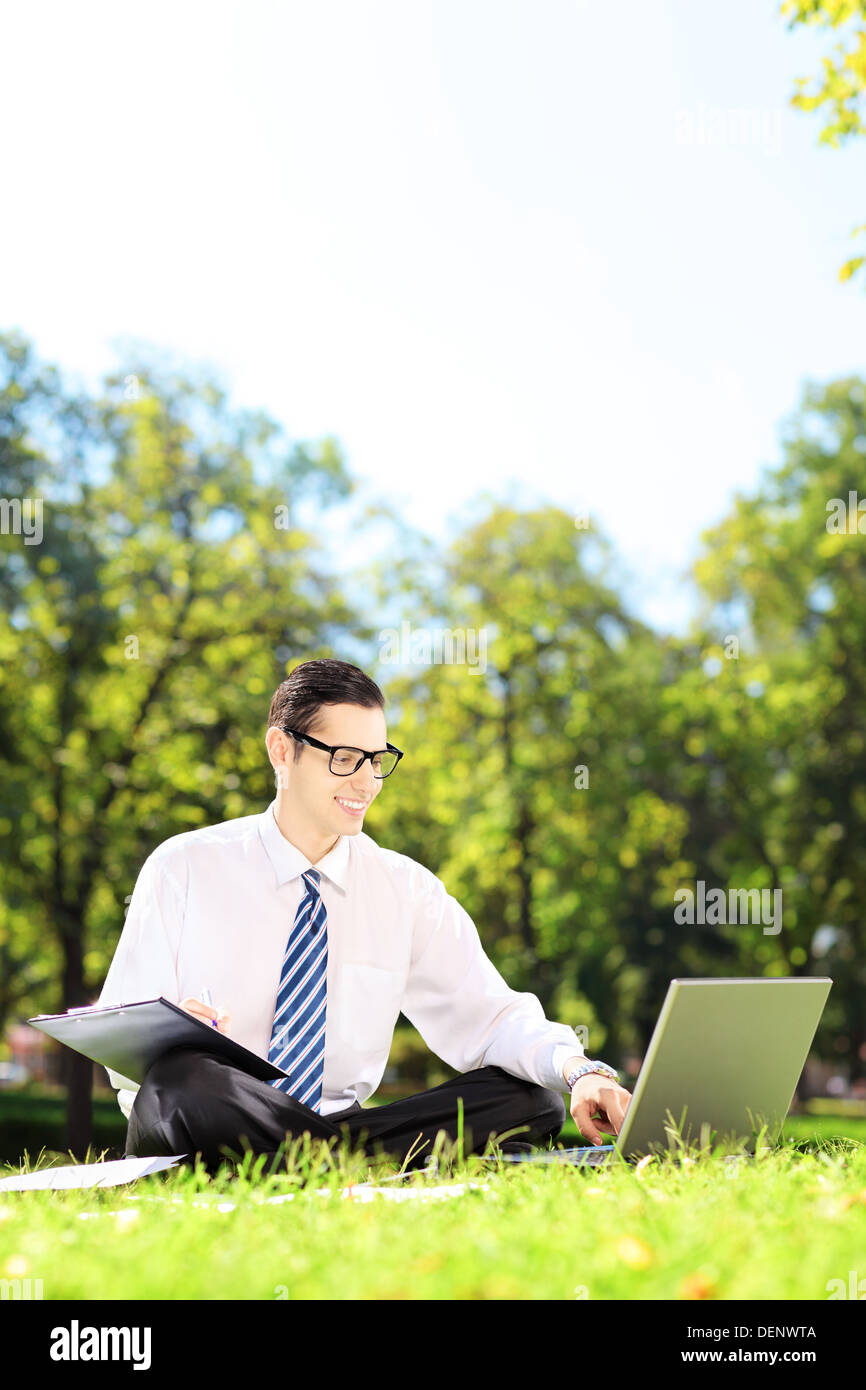 Junger Geschäftsmann sitzt auf einer Wiese auf einem Laptop arbeiten und aufschreiben von Notizen in einem park Stockfoto
