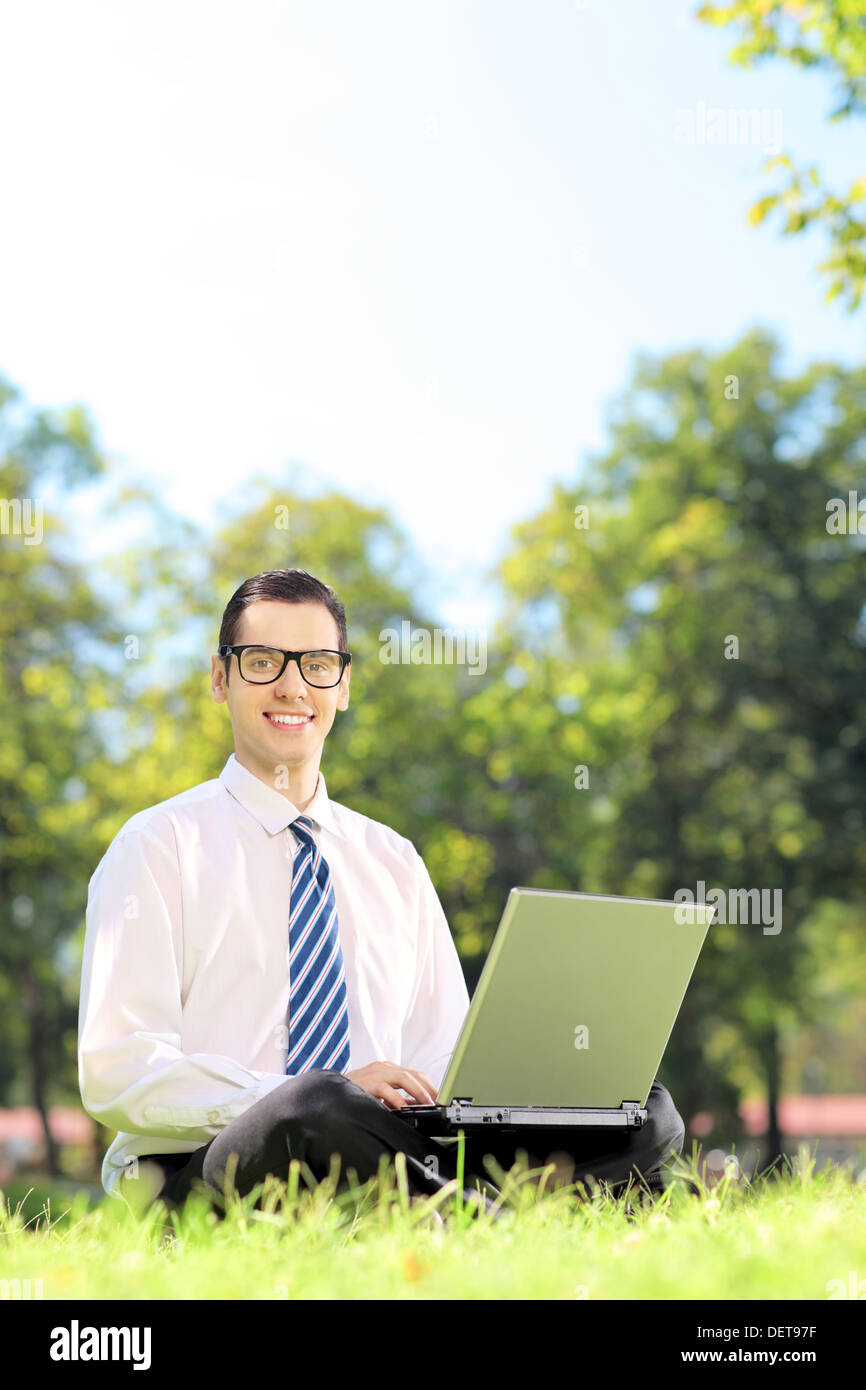 Lächelnd jungen Geschäftsmann sitzt auf einer Wiese arbeitet auf einem Laptop in einem park Stockfoto