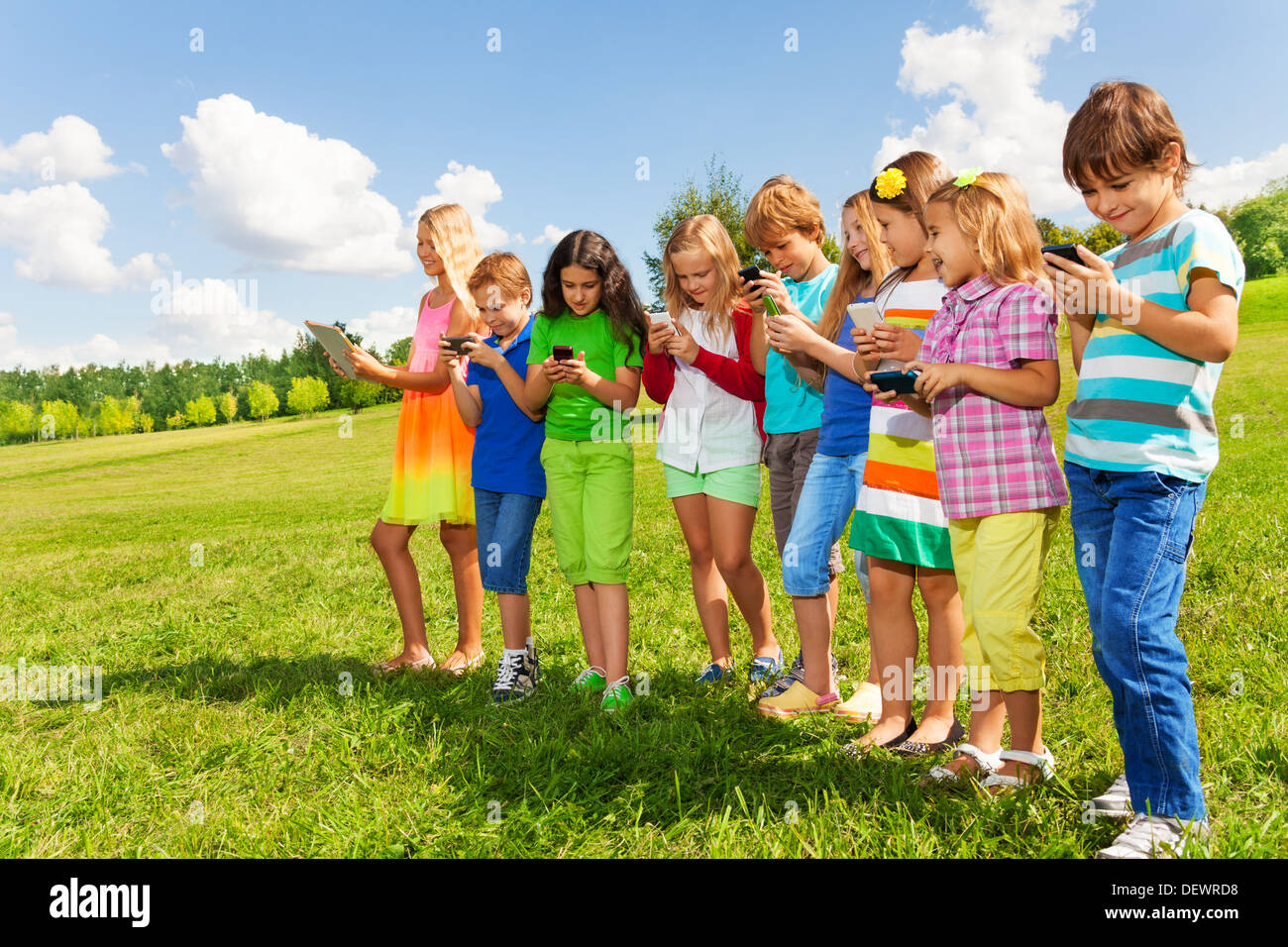 Große Gruppe beschäftigt Kinder, Jungen und Mädchen auf ihren Handys sms sms und Spielen im Freien im Bereich bleiben auf hellen, sonnigen Sommer Tag suchen Stockfoto