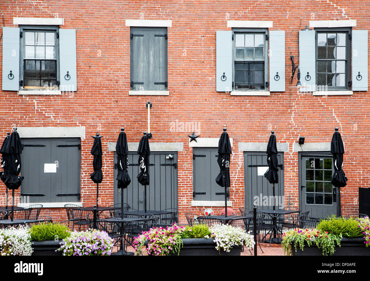 Eine alte Backsteingebäude mit grauen Türen und Fensterläden und geschlossenen schwarzen Schirmen von Tabellen auf Terrasse mit Blumen in den Blumenkästen Stockfoto