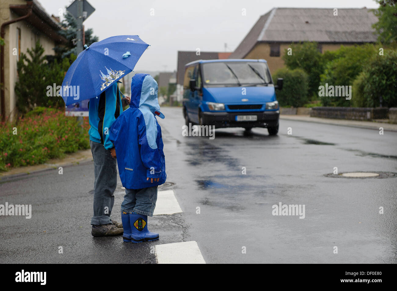 Zwei Kinder, 4 und 8 Jahren darauf warten, überqueren Sie die Straße im Regen, während ein Auto, Assamstadt, Baden-Württemberg nähert Stockfoto