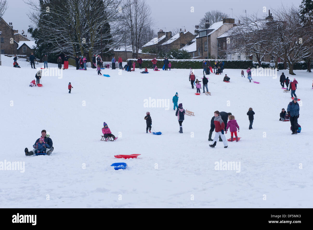 Viele Personen (Erwachsene und Kinder) mit Spaß für die ganze Familie im Winter Schnee, Rodeln, Down Hill in Parkstellung - Riverside Gardens Ilkley, Yorkshire, England, UK. Stockfoto