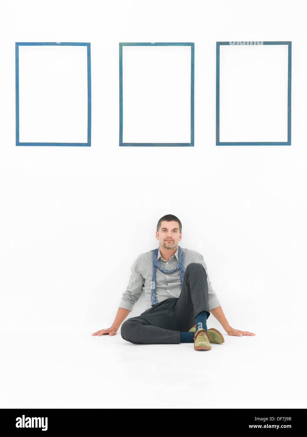 Vorderansicht der Mann sitzt auf dem Boden in eine entspannte Haltung gegen eine weiße Wand mit drei leeren blauen Rähmchen Stockfoto