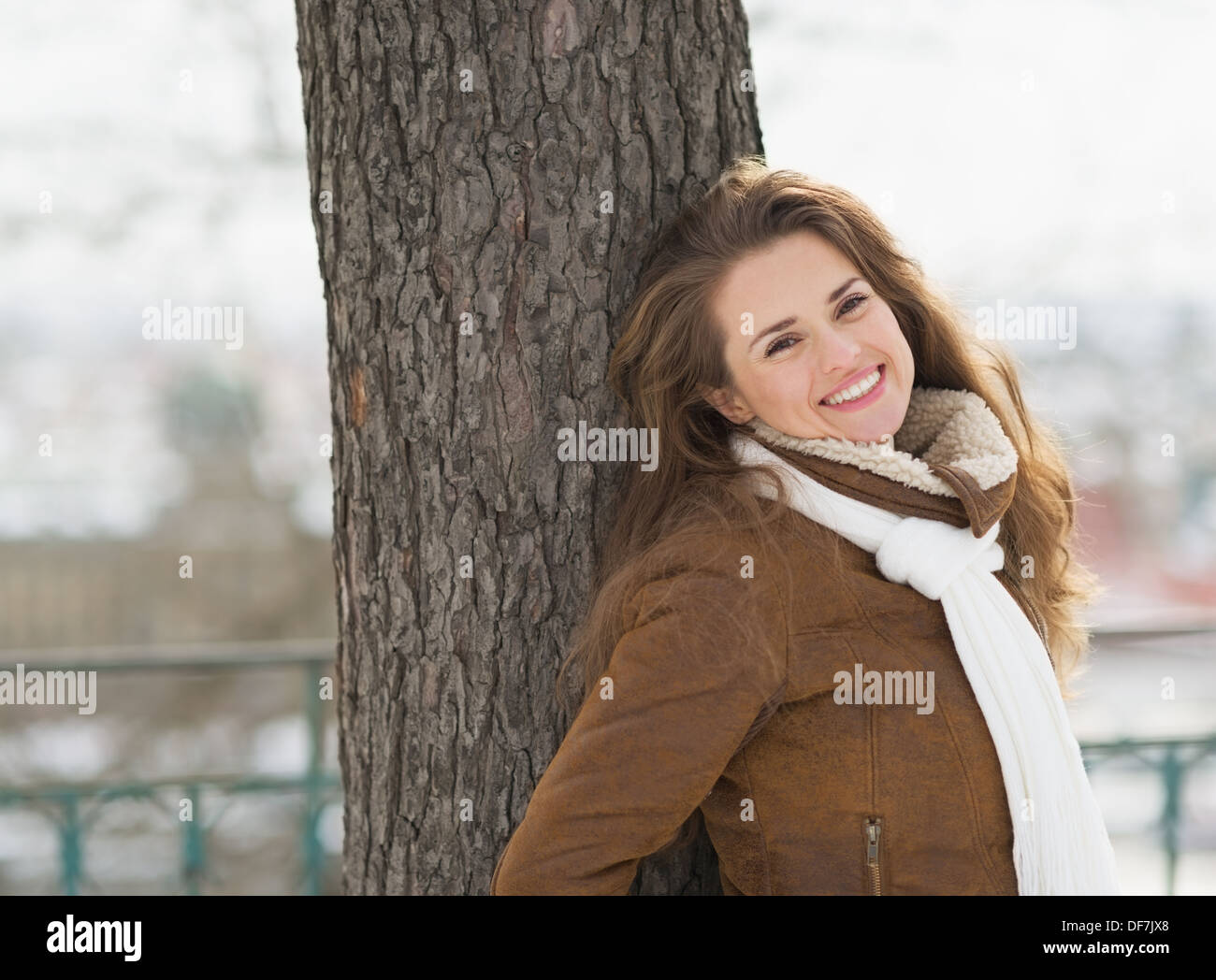 Lächelnde junge Frau lehnte sich gegen Baum im Winter park Stockfoto