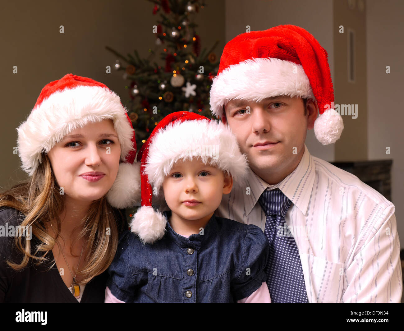 Porträt von Mama, Papa und ihre süße kleine Tochter alle tragen Weihnachtsmann Mützen mit Weihnachtsbaum im Hintergrund Stockfoto