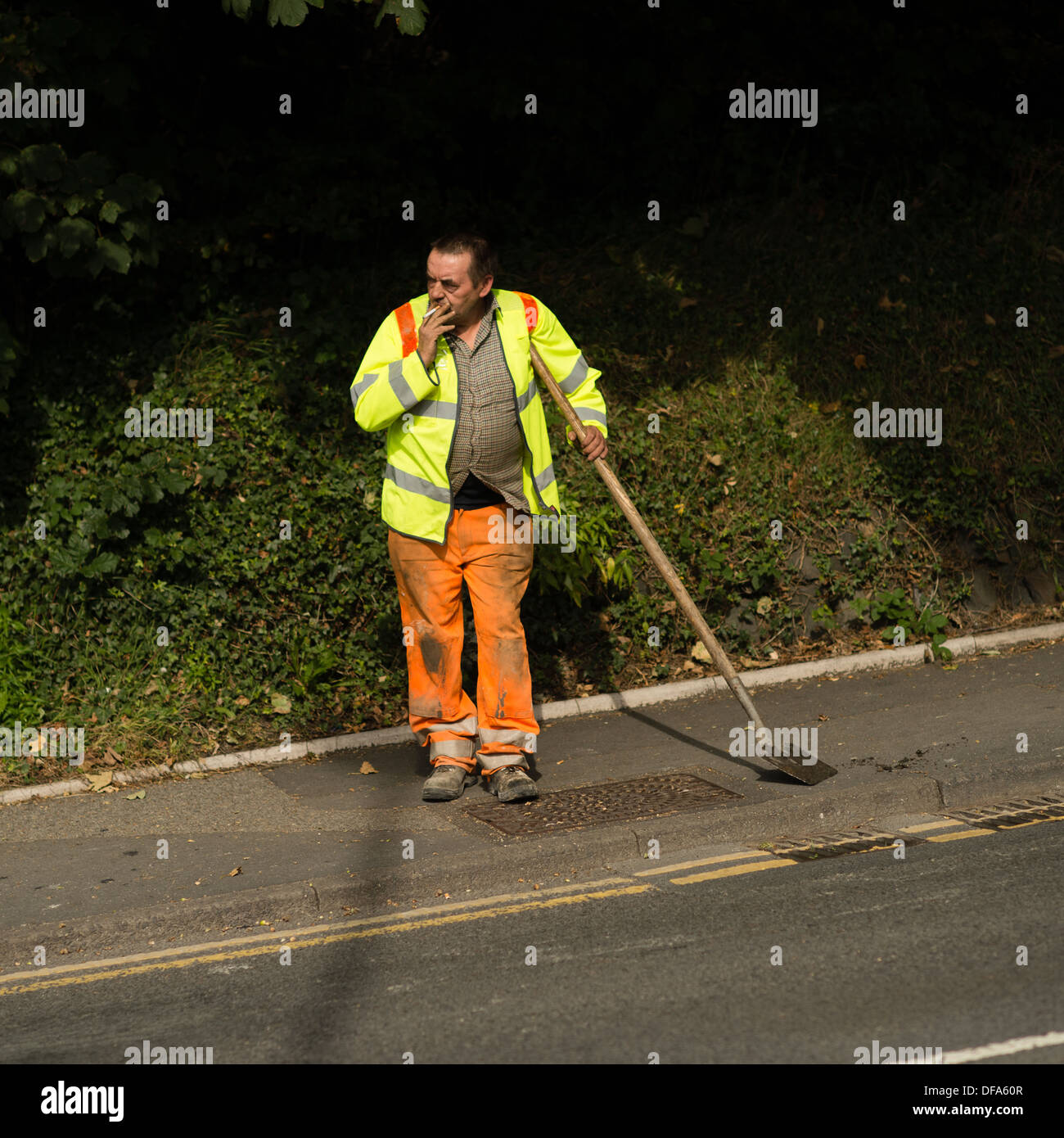 Ein kommunalen Rat direkte Arbeit Arbeiter orange Hose und eine helle gelbe hi-Vis Warnschutz Jacke eine Pause von seiner Arbeit und stützte sich auf seinen Spaten Schaufel Rauchen einer Zigarette UK Stockfoto