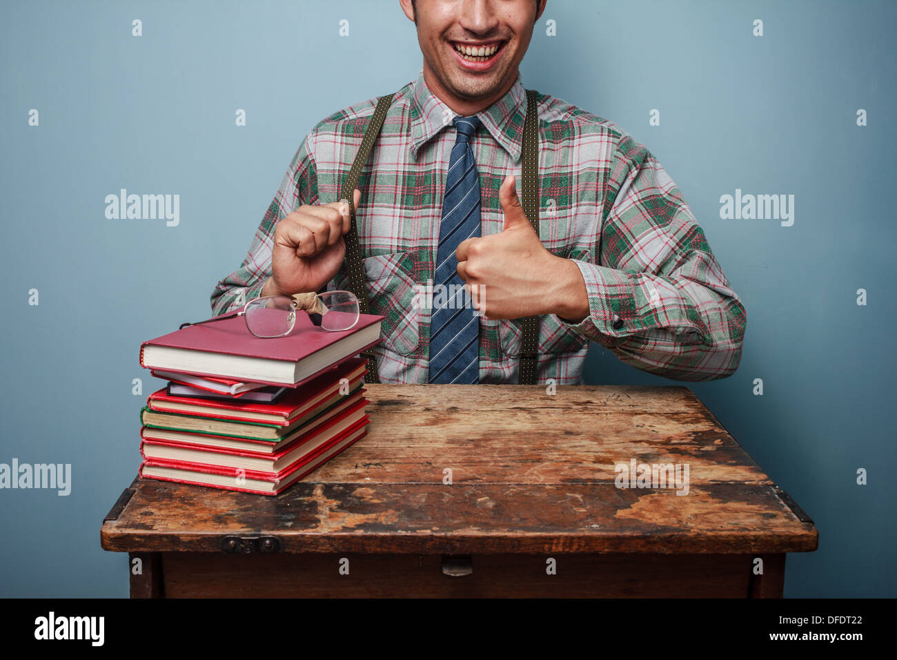 Jungen Nerd Daumen aufzugeben, da er neben einem Stapel Bücher sitzt Stockfoto