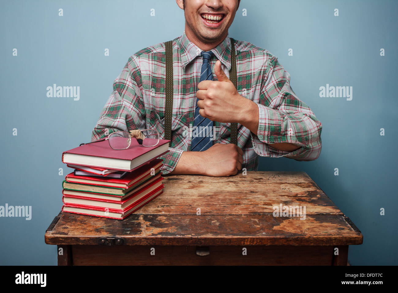 Jungen Nerd Daumen aufzugeben, da er neben einem Stapel Bücher sitzt Stockfoto