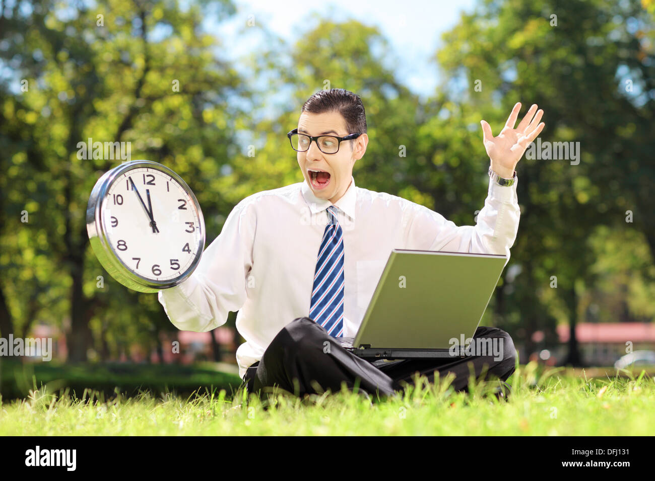 Junge wütend Unternehmer mit Laptop auf dem Rasen sitzen und Blick auf die Uhr in einem park Stockfoto