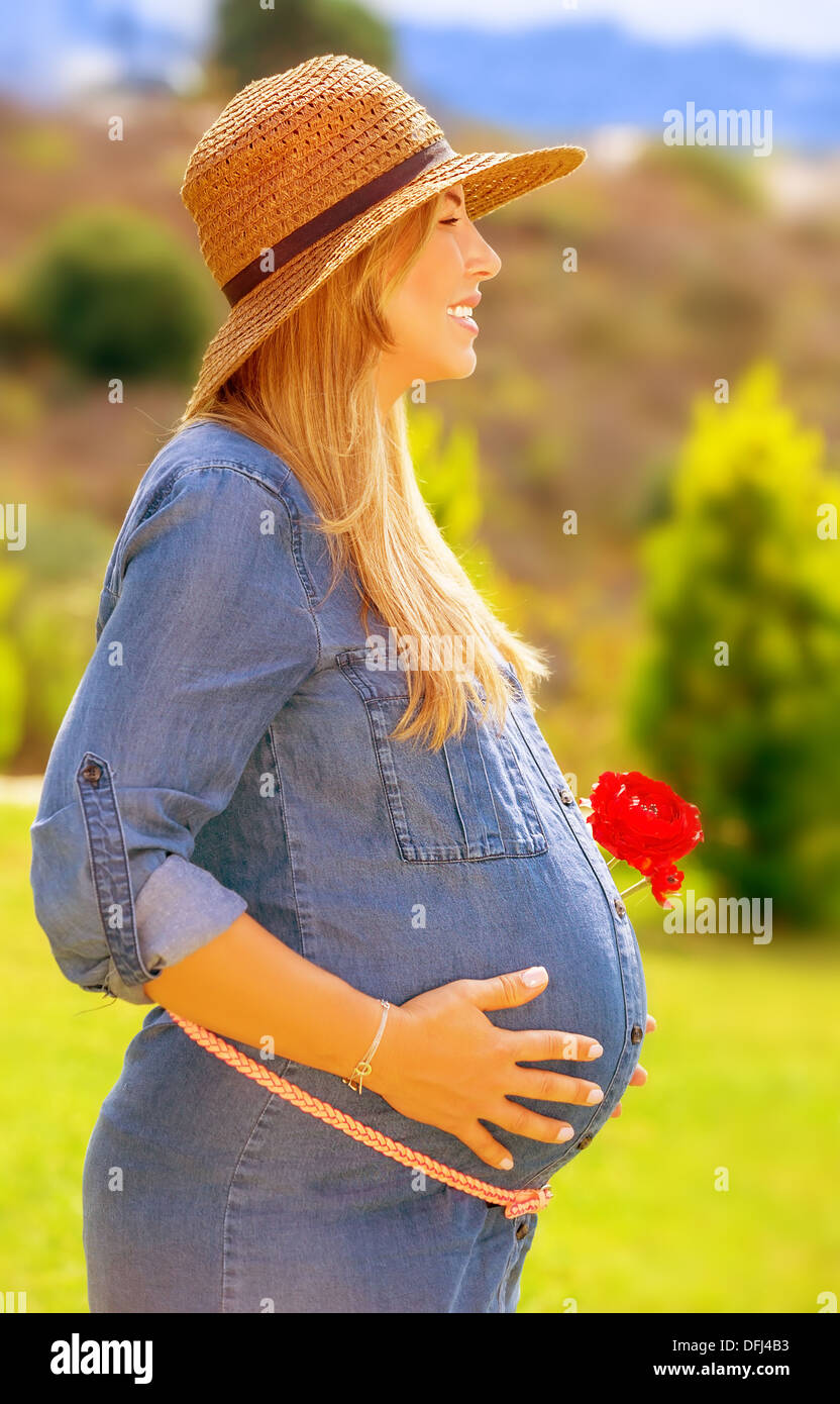 Schwangere Frau im Park, Seitenansicht, sonnigen Tag, rote blumeblumenstrauß, glückliche Schwangerschaft, berühren, Bauch, Liebe und neues Leben Stockfoto