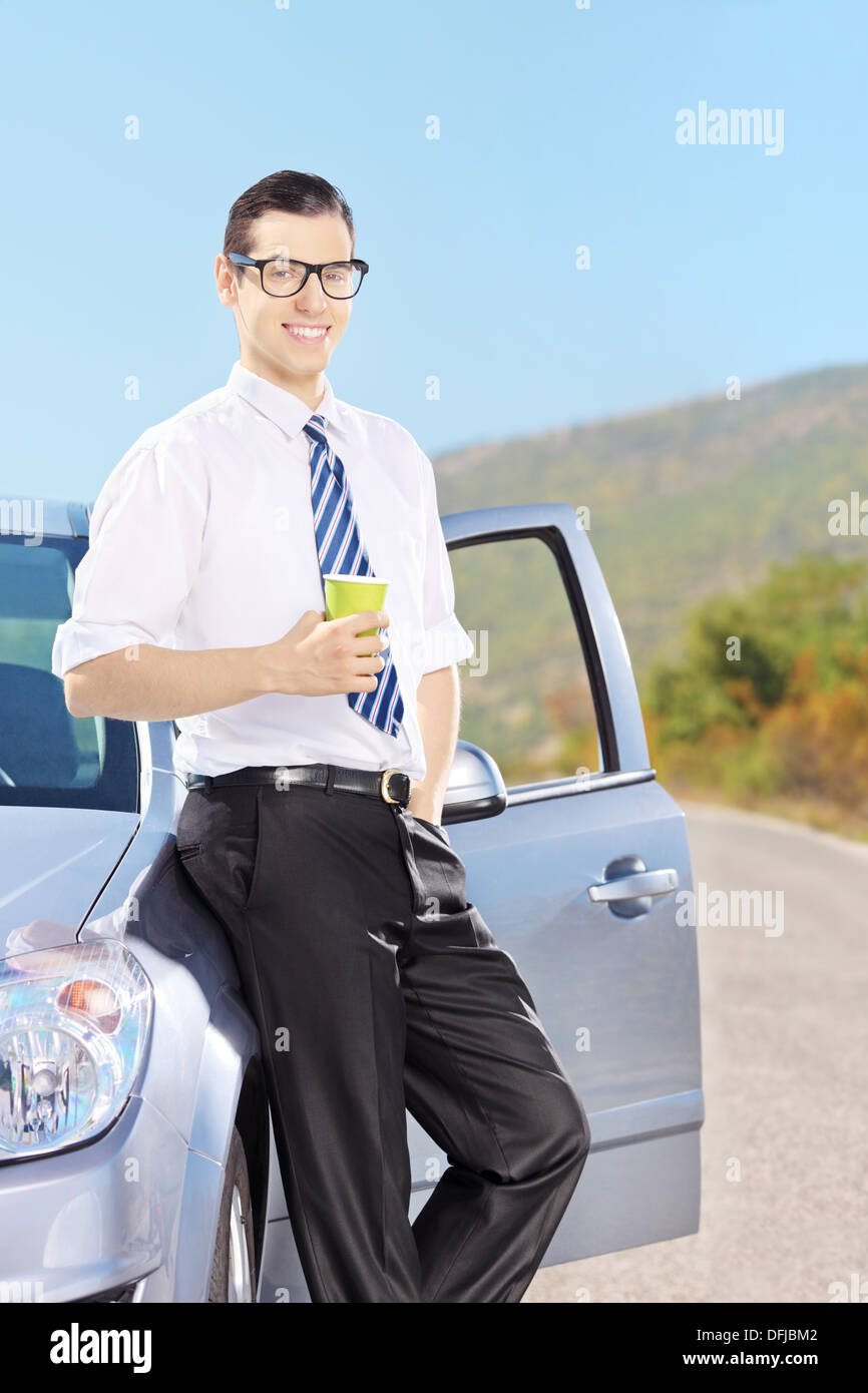 Lächelnde junge Mann auf seinem Auto hält eine Kaffeetasse auf einer Straße Stockfoto