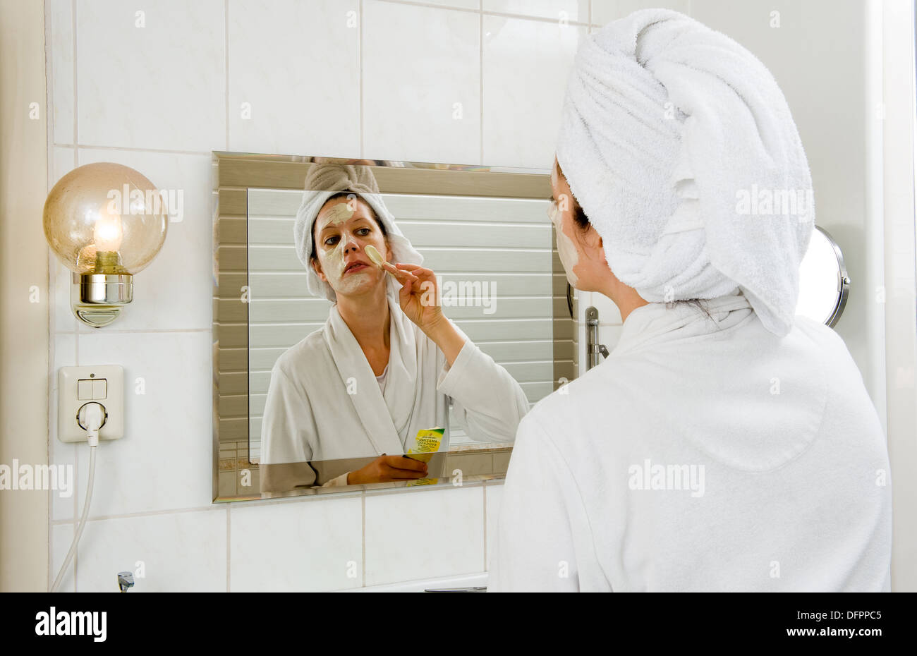 Eine Frau ist eine Maske auf ihr Gesicht vor dem Spiegel im Badezimmer verschmiert. Stockfoto