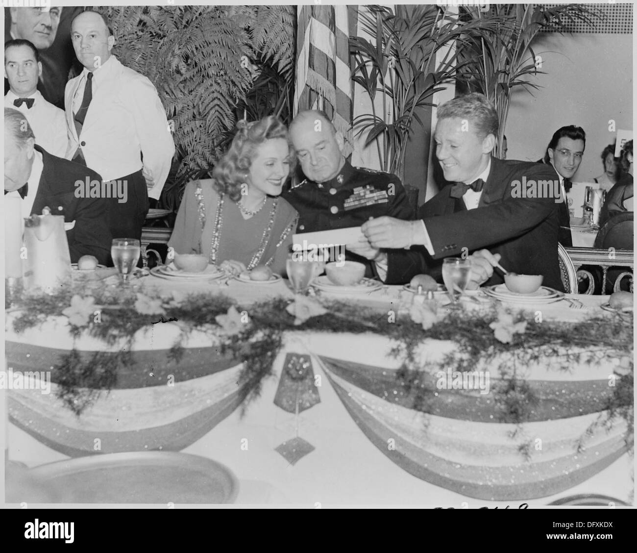 Dieses Foto zeigt die Filmstars Constance Moore und Van Johnson, die mit einem nicht identifizierten Militäroffizier an einem Tisch sitzen. Das Bild spiegelt die Unterhaltungsindustrie Mitte des 20. Jahrhunderts wider. Stockfoto