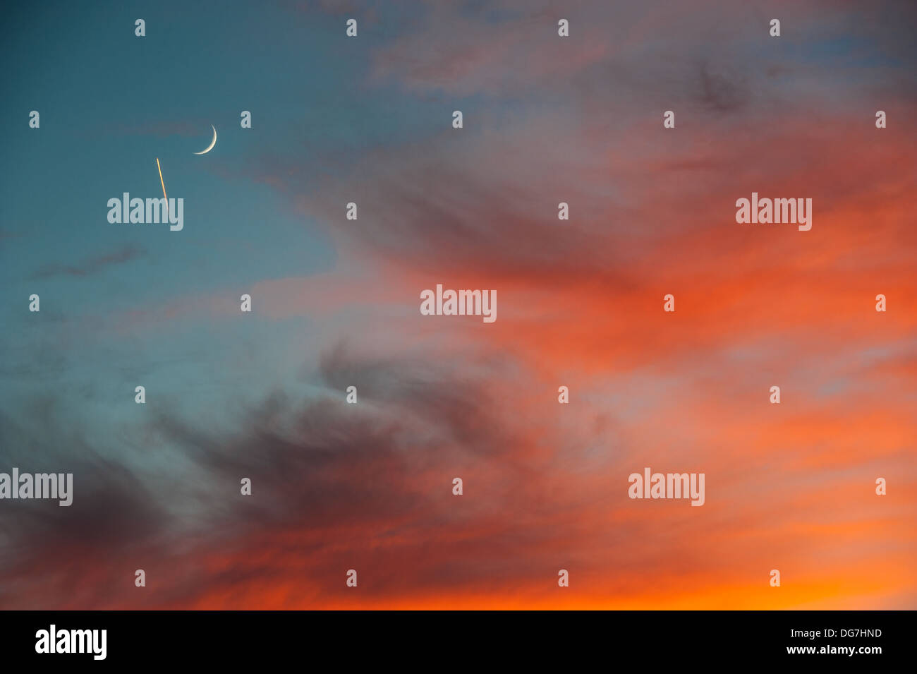 Halbmond und Flugzeugjet kontrastieren in einem feurigen Himmel von tiefblauem und orangefarbenem Sonnenuntergang. (USA) Stockfoto