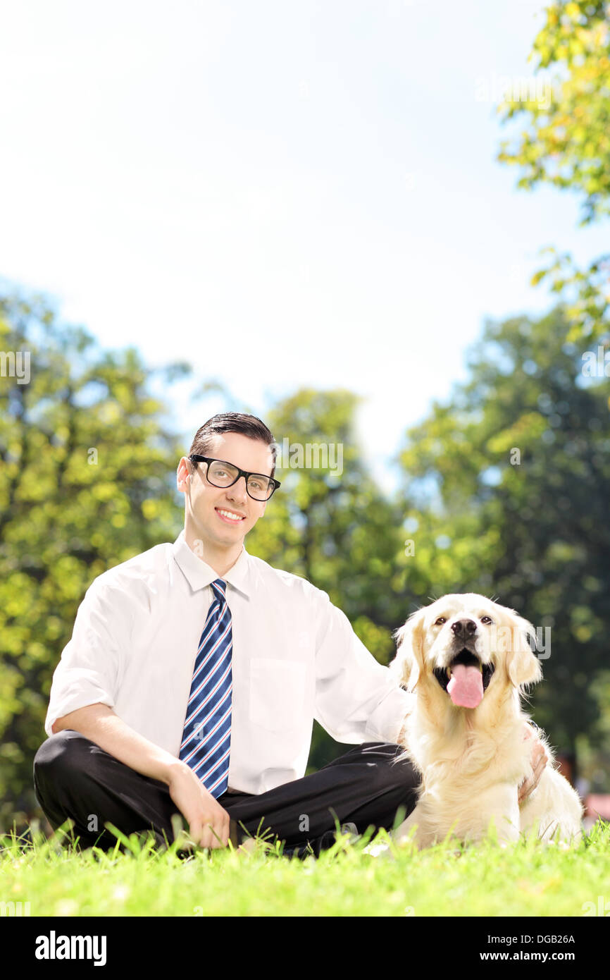 Junger lächelnder Mann sitzt auf einem grünen Rasen neben seinem Hund in einem park Stockfoto