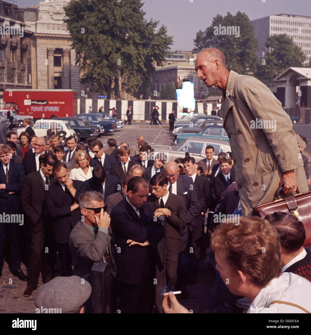 1960er Jahre, historisch, sommerlich und ein Mann mit Regenmantel und Lederaktentasche, ein Seifenboxen-Lautsprecher, spricht vor einer Menschenmenge am Mittag im Tower Hill, London, EC3 England, UK. Tower Hill und die Rednerecke im Hyde Park waren die beiden beliebten Orte in London für freie Meinungsäußerung unter freiem Himmel. Stockfoto