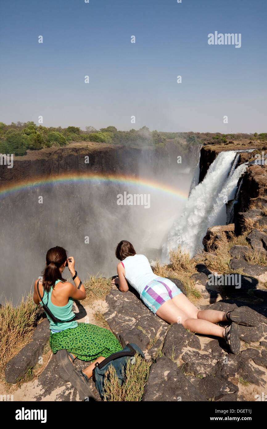 Zwei Frauen suchen über den Rand an den Victoria Wasserfällen mit Regenbogen, Sambia Seite Insel Livingstone, Sambia Afrika Stockfoto