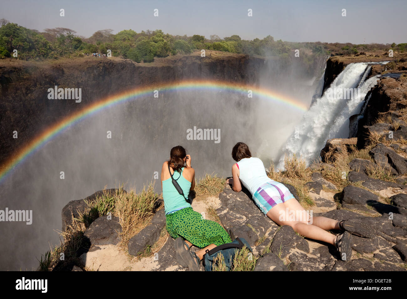 Victoria Falls Sambia; Zwei Touristen für Frauen, die mit Regenbogen, Sambia-Seite, Livingstone Island, Sambia-Afrika über den Rand der Victoriafälle blicken Stockfoto